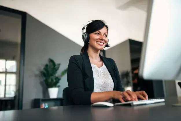 A professional call center agent smiling while providing outsourced contact centre services through a desktop computer, wearing a headset in a modern office environment.