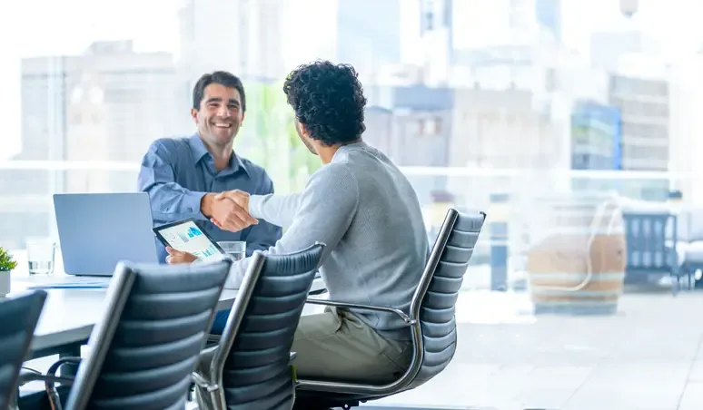 Two businessmen shaking hands in a modern office boardroom after closing a deal, with a digital tablet and laptop in front of them—symbolizing successful call ring support collaboration.
