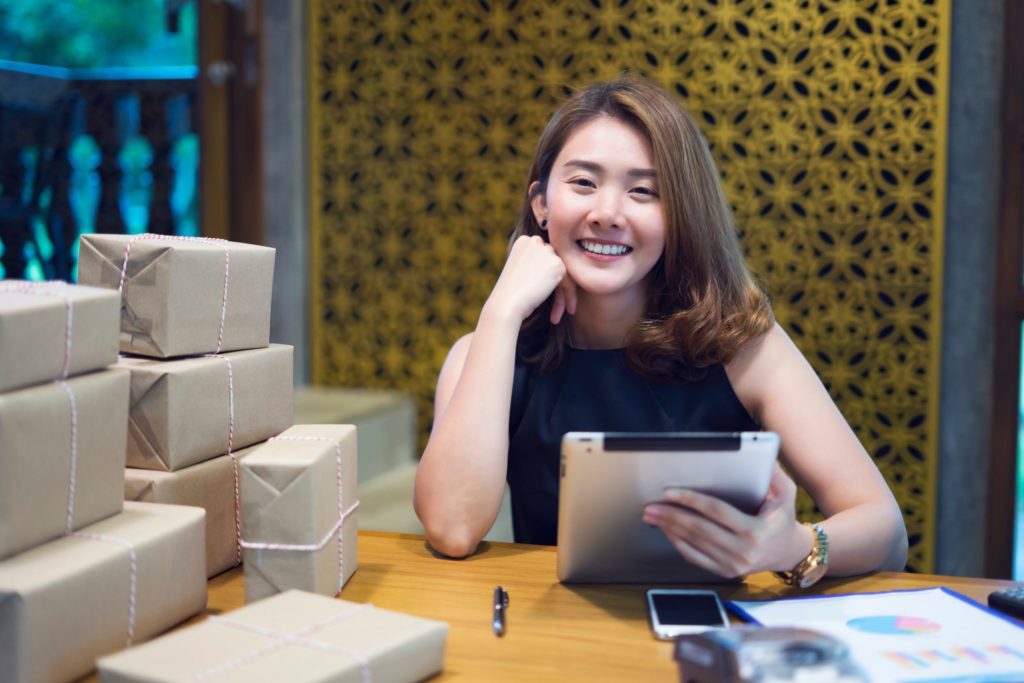 A content and happy Vietnamese woman is smiling while browsing her iPad tablet because she ordered many eCommerce packages from an eCommerce marketplace.