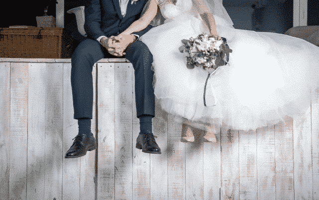 A beautifully dressed bride in a fluffy white wedding dress is grabbing the handsome dressed-in-a-blue-suit groom's hand while also leaning on his shoulder while they both sit on a vintage white-painted fence; in the bride's right hand is a bouquet of white freshly-bloomed flowers.