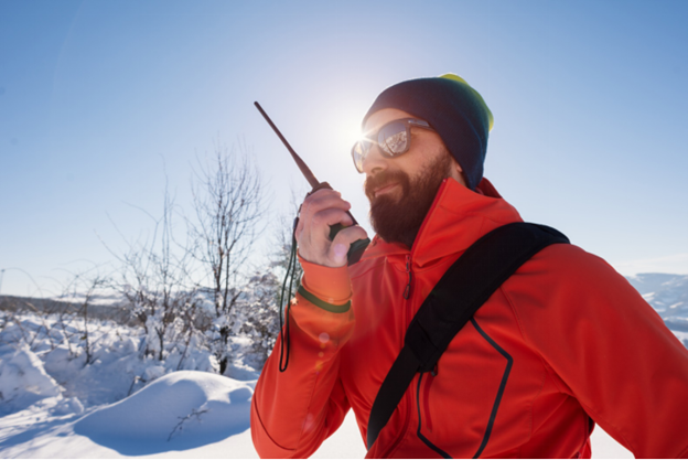 A mountain climber dressed in a red winter coat and a black winter hat is calling his colleague who is found at a higher point of the mountain to coordinate his scale up the mountain; it is winter time.
