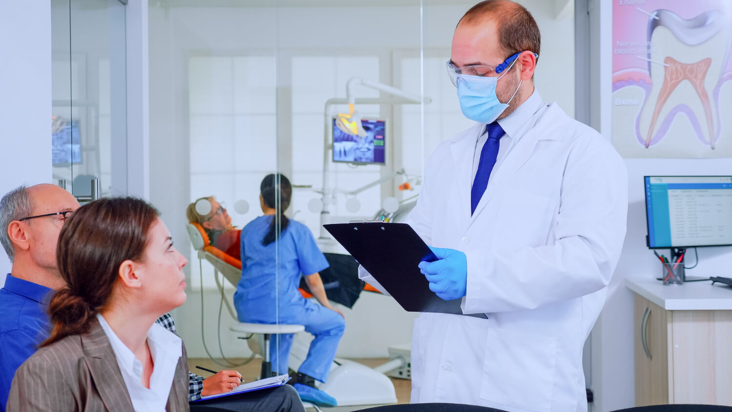 A dentist carefully-reviews with the wife of the man receiving dental care via a dental assistant in the dental room in the background his current dental situation and what treatment options are available; the dentist takes care to follow dental office HIPAA compliance best practices while sharing such patient data.