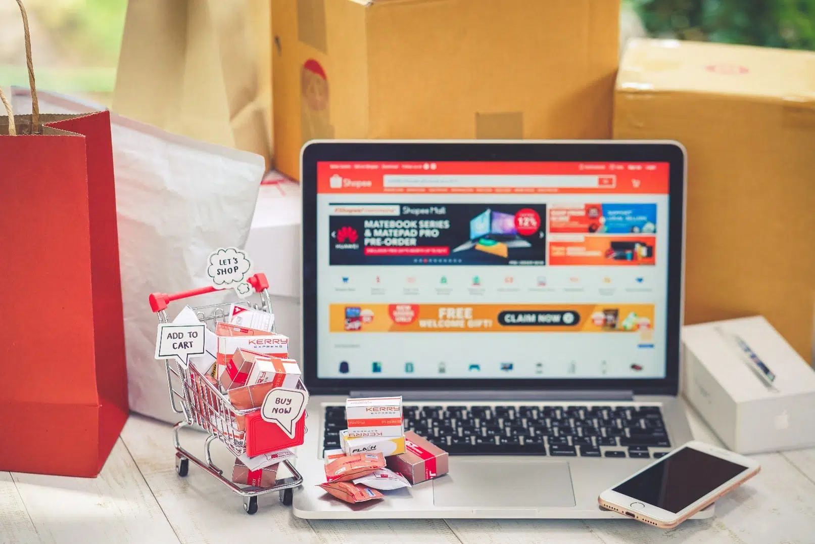 A computer is showing the Shopee Malaysia eCommerce market website while a shopping cart next to it says, "Let's Shop," "Add to Cart," and "Buy Now;" there is a shopping bag in the picture, a newly-purchased iPhone, and the shopping cart is filled with eCommerce packages.