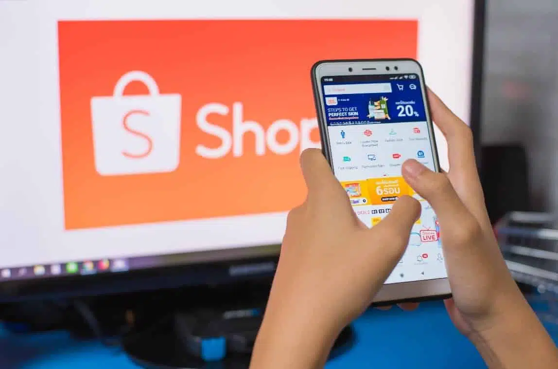 A Thai eCommeerce consumer is shopping on their cellphone for products on Shopee Thailand's e-Commerce platform; there is a Shopee logo in the background on a computer monitor.